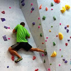 Man in green shirt and black shorts rock climbing on an indoor wall with colorful holds.