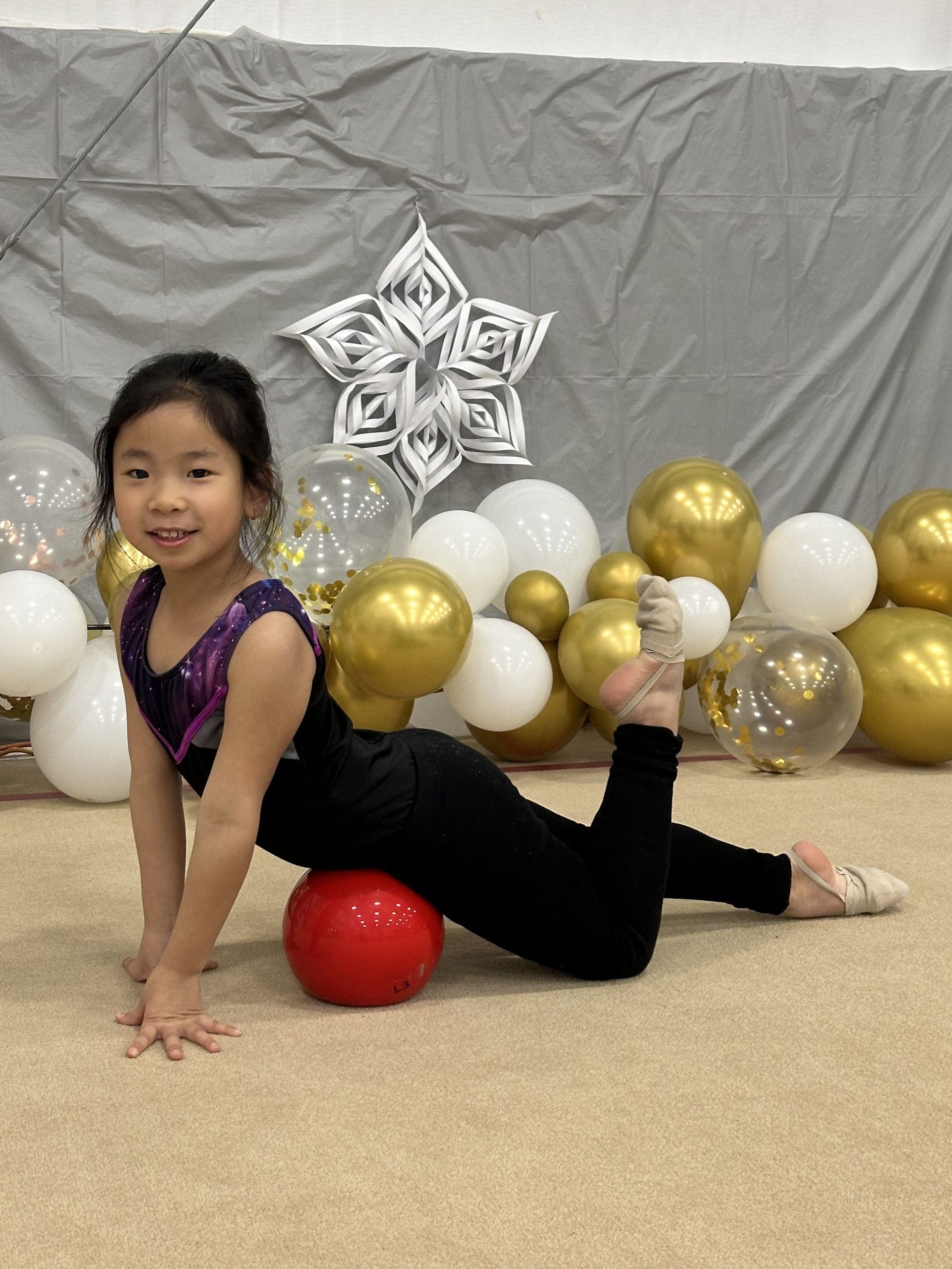 Young gymnast in black leotard poses with a red ball, smiling, surrounded by balloons, indoors.