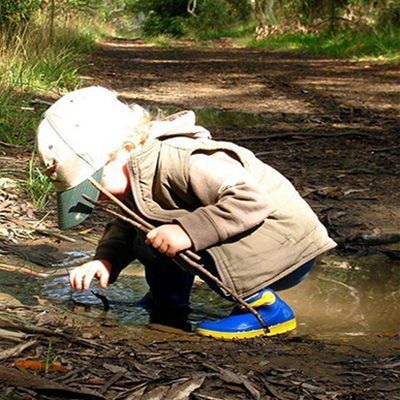 Child in boots playing in muddy puddle, holding sticks, in a wooded area.