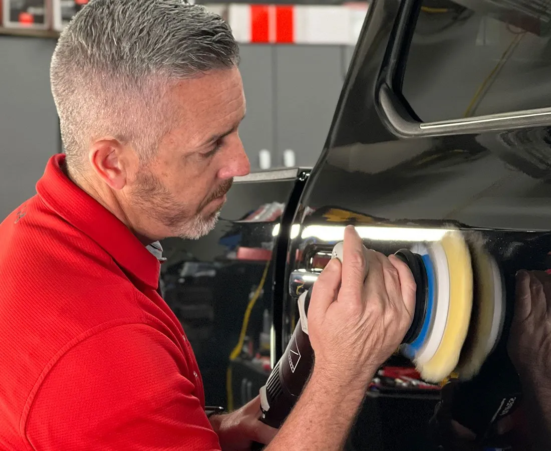 Man in red shirt polishing a black car with a rotary buffer in a workshop.