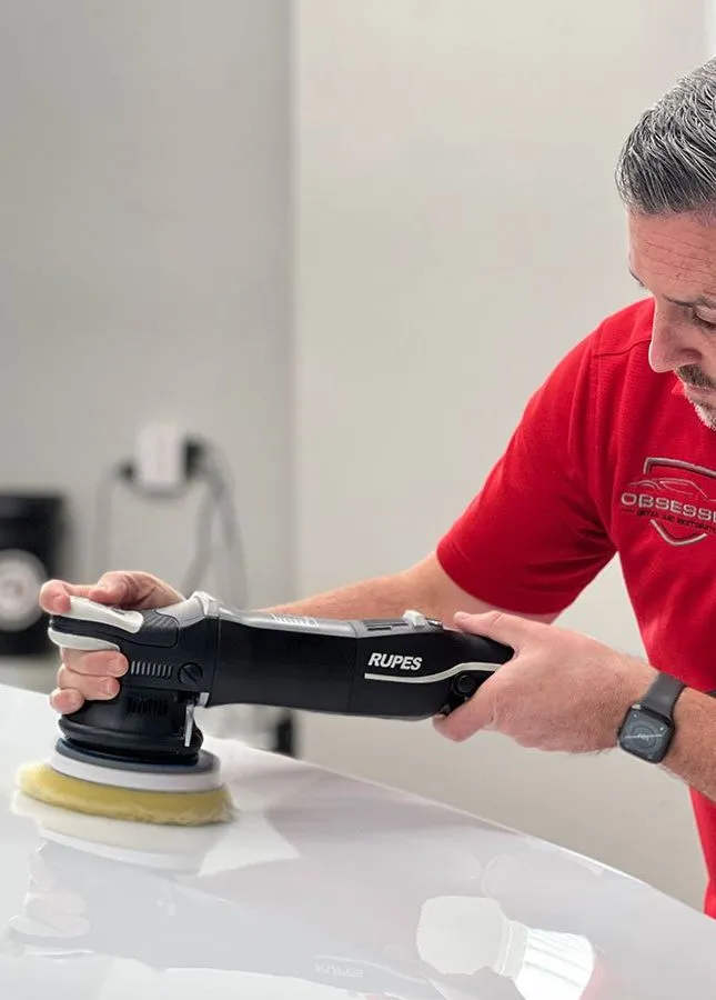 Man in red shirt polishes a white car with a black buffer in a white-walled room.