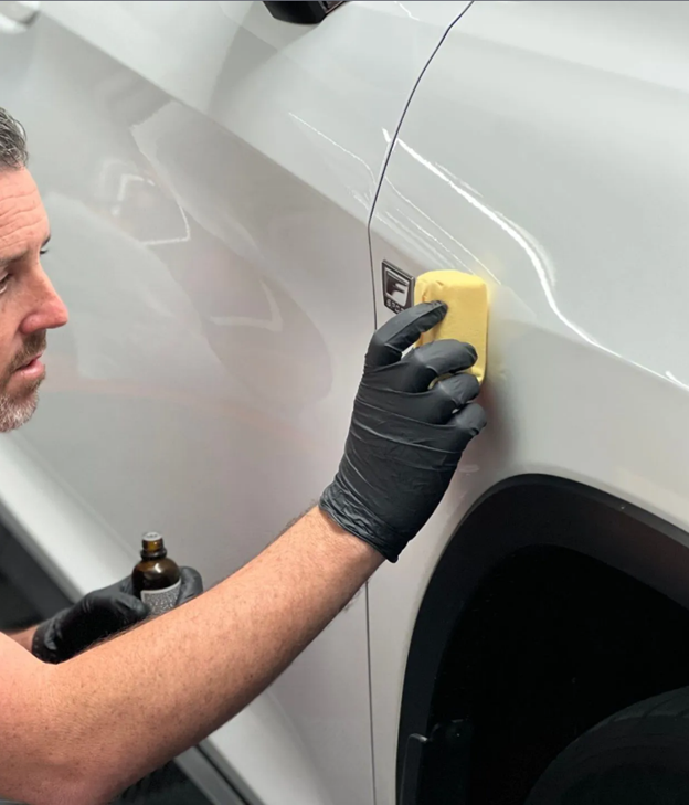 Man in black gloves applying a coating to a white car panel with a yellow applicator.