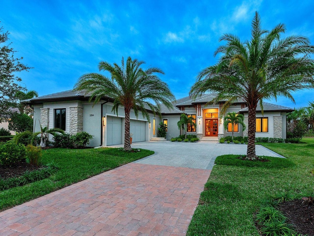A modern, light-colored single-story home with a brick driveway and palm trees against a twilight sky.