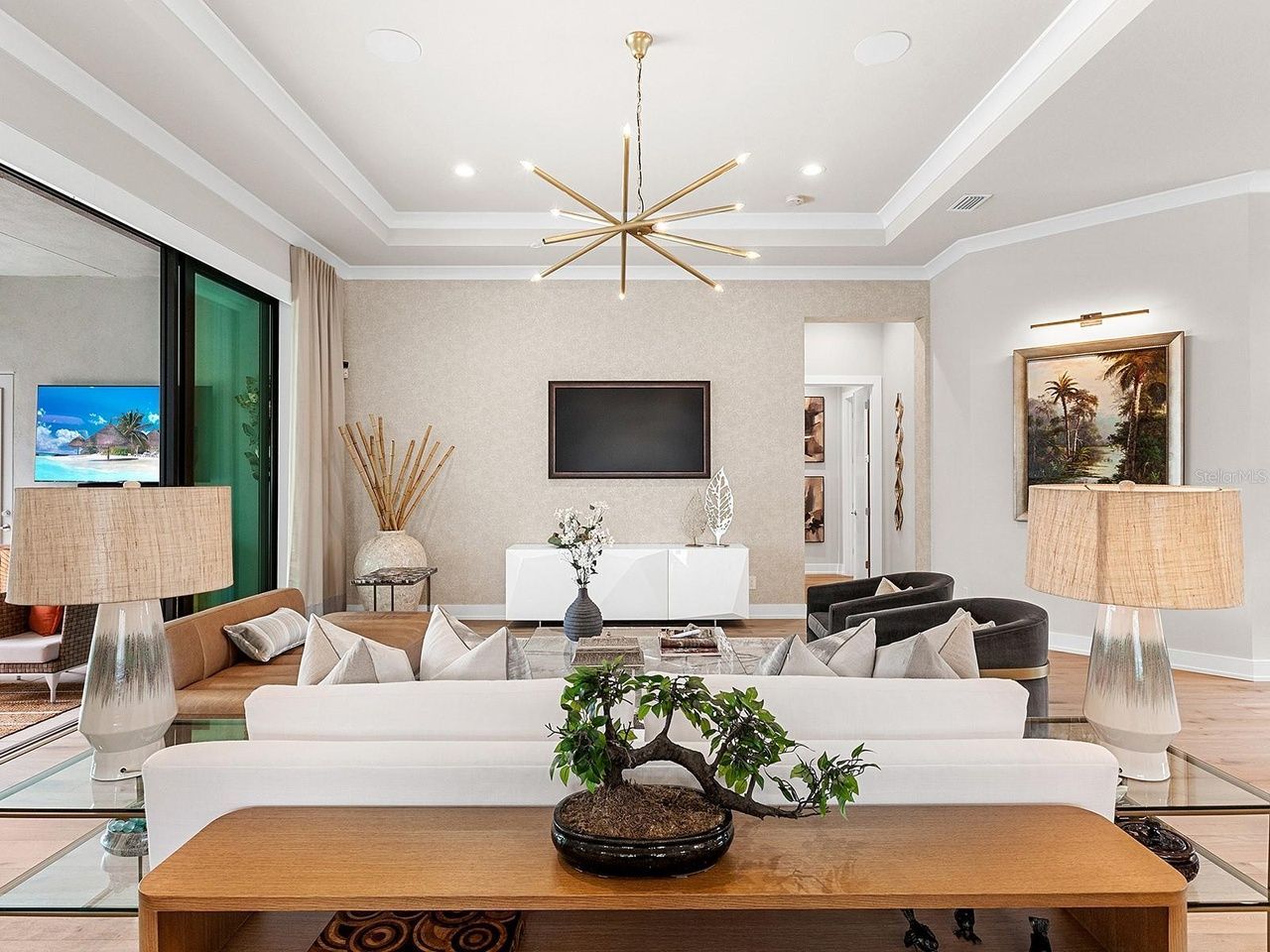 A modern, neutral-toned living room featuring a white sofa, bonsai plant, gold chandelier, and a large wall-mounted TV.
