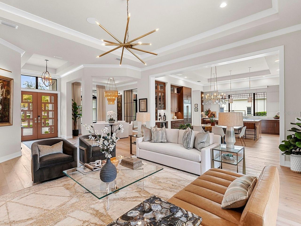 An open-concept living area with a modern gold chandelier, beige sofa, dark armchair, and a kitchen visible in the back.