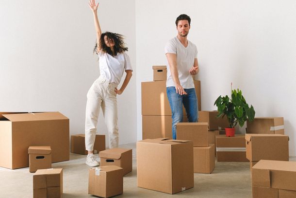 Two people among moving boxes in a bright room, one waving and one standing beside a plant.