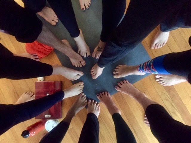 Woman smiling in a yoga pose on a wood floor. Others in the background.