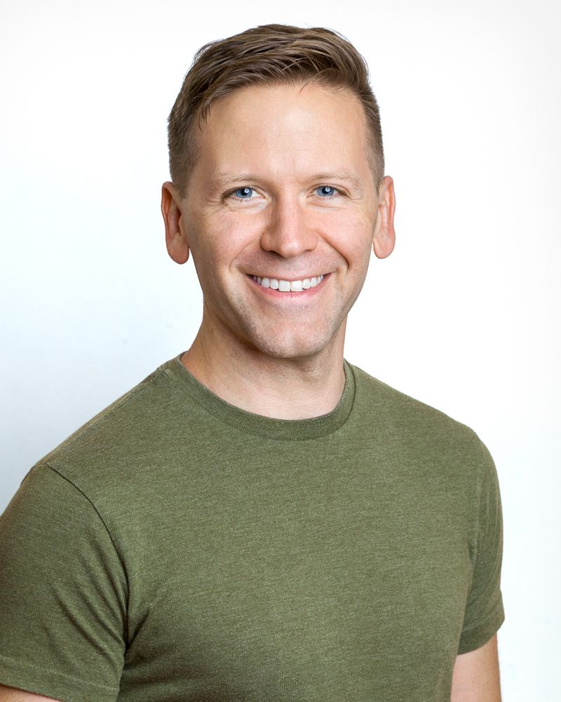 Smiling man with short brown hair and blue eyes in a gray shirt against a blue background.