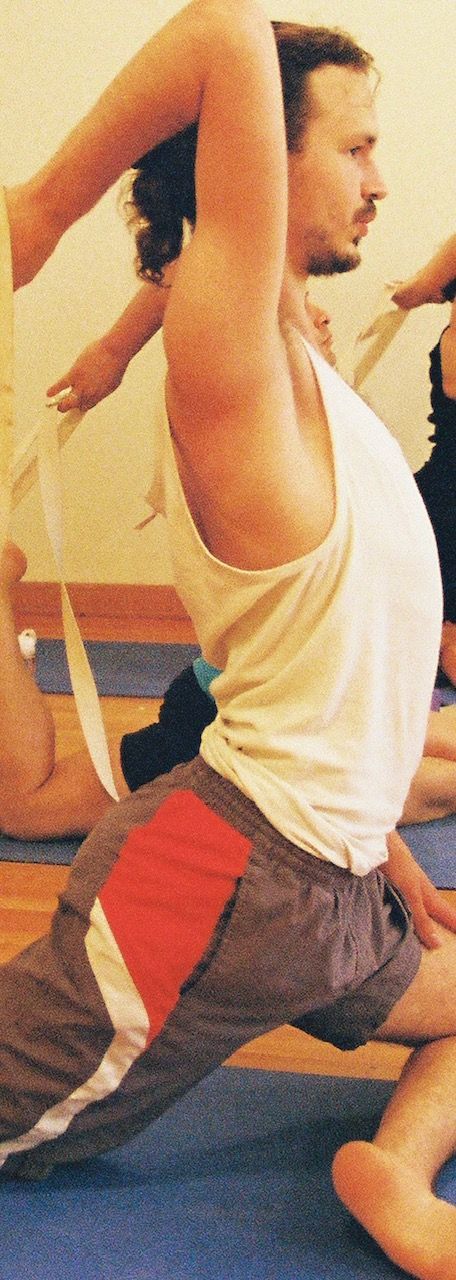 Man in yoga pose, arm raised, side profile. Wearing tank top, shorts with red panel. Blue mat, group setting.