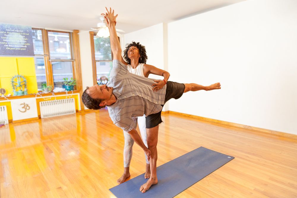 Woman doing yoga on a stone wall outdoors with view of water; wearing light jacket, patterned pants, and a tank top.