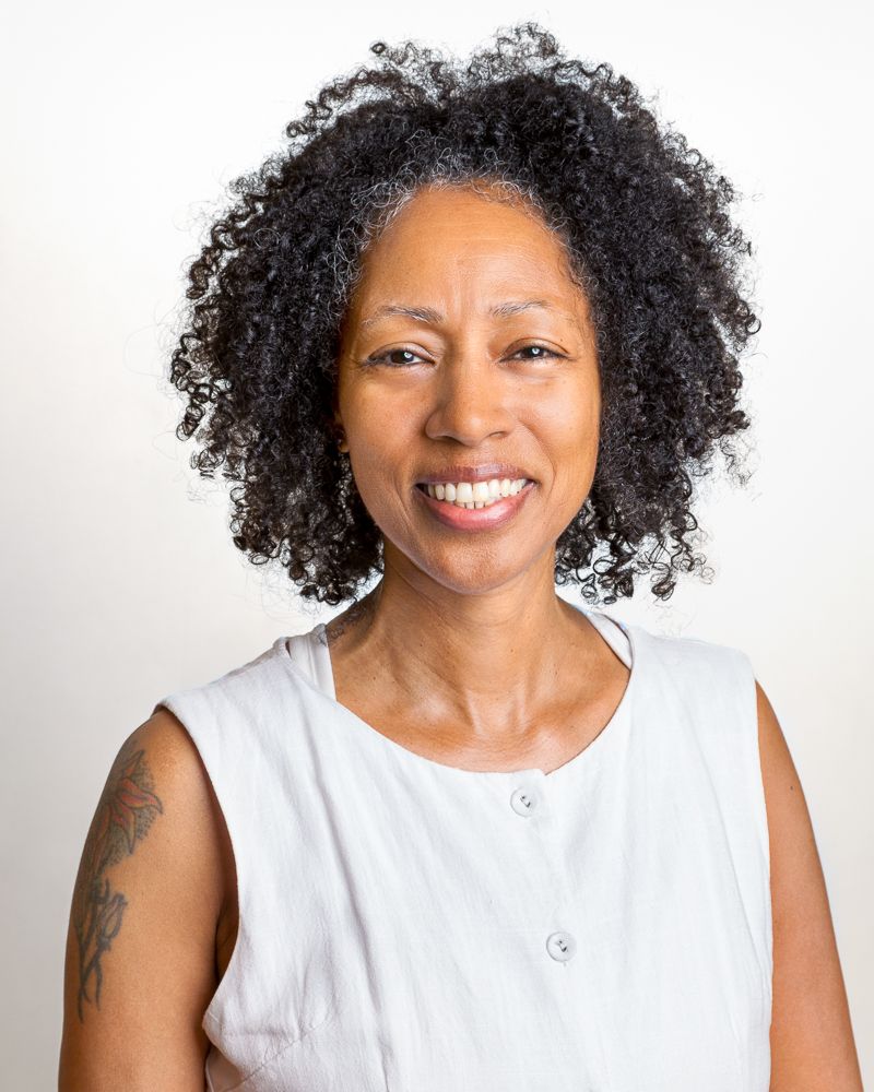 Woman doing yoga on a stone wall outdoors with view of water; wearing light jacket, patterned pants, and a tank top.