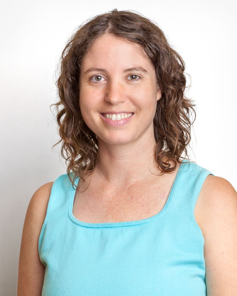 Woman with brown curly hair smiles at the camera in front of a blurry forest background.