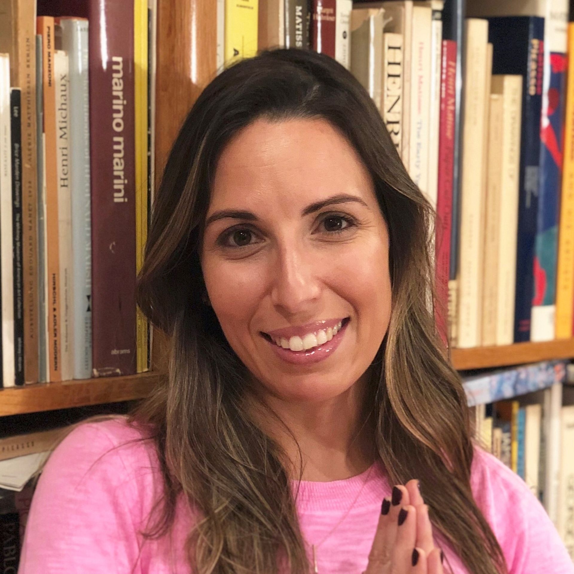 Brunette woman with an inviting smile stands in front of a bookshelf with her hands in pranam.