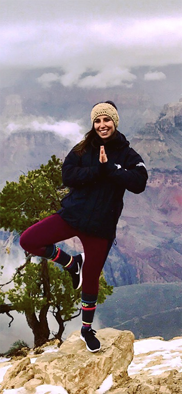 Woman doing a tree pose on a rock at the Grand Canyon, smiling, wearing a hat, dark coat, and burgundy leggings.