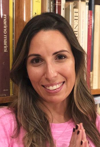 Woman with long brown hair, smiling in front of a bookshelf, wearing a pink shirt.