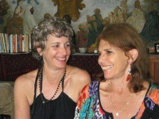Two smiling women, one in black halter top, other in colorful print, near a shelf of books, with a painted backdrop.
