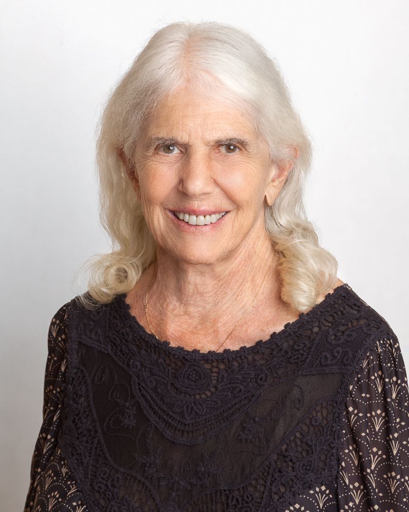 Smiling woman with long brown hair and blue top, wearing earrings and a necklace, against a light background.