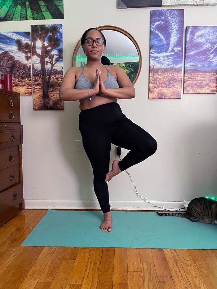 Woman in yoga pose, on one leg, hands in prayer, wearing gray top and black leggings, on a teal mat indoors.