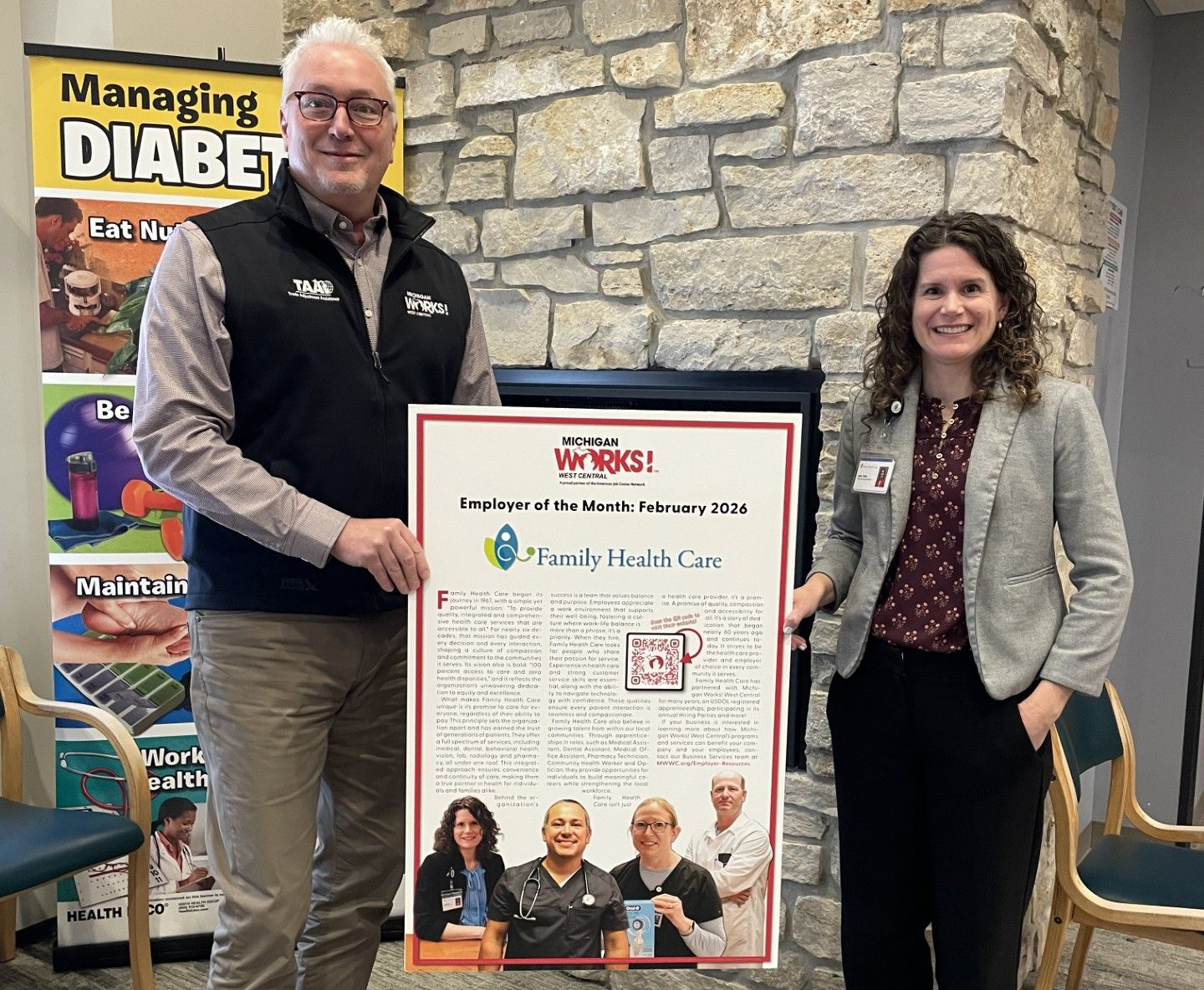 Two people holding a poster: managing diabetes info. Man wears a vest, woman a blazer, near a stone wall.