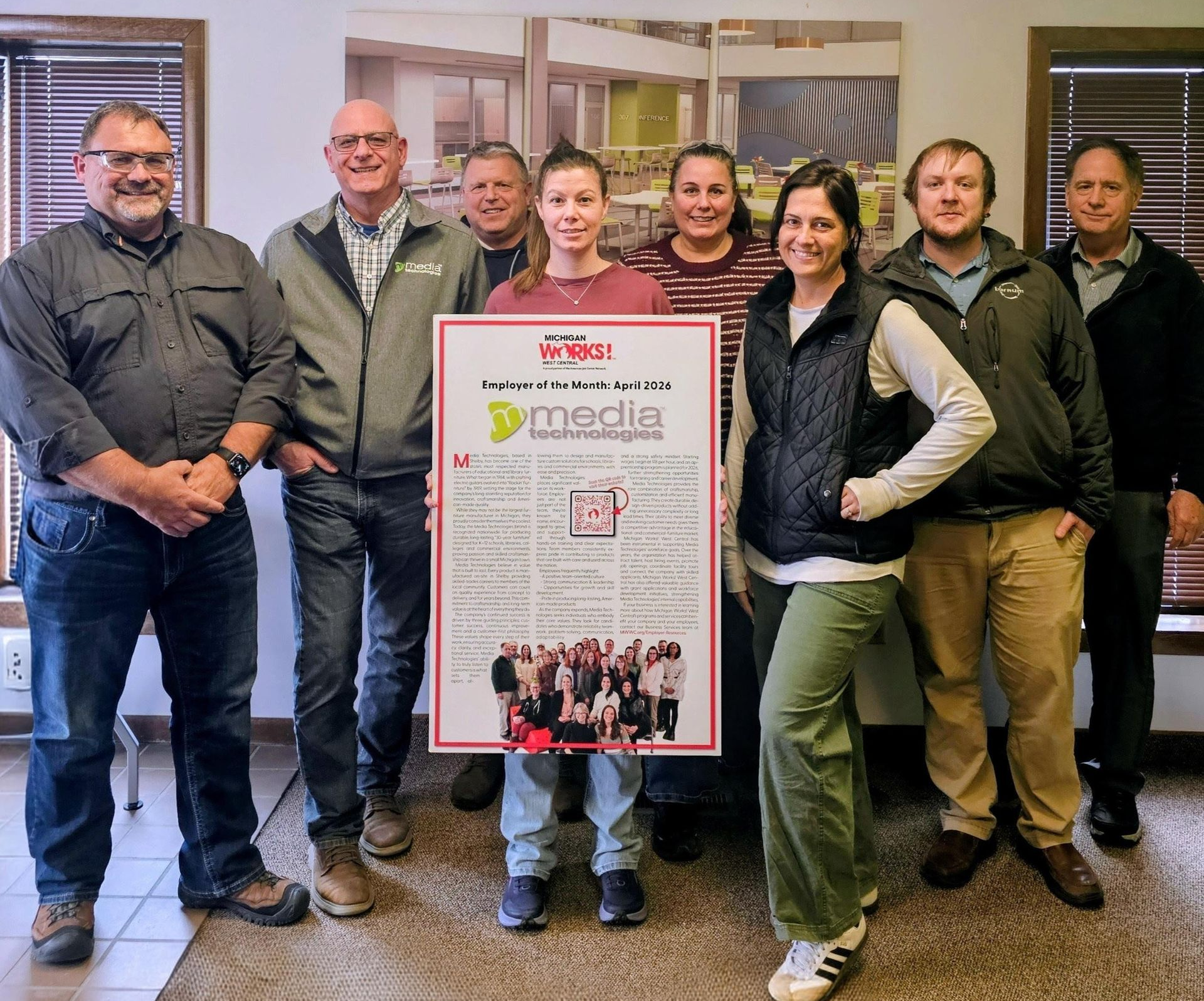 A group of eight people stand in an office, holding a poster with a company logo and a team photo for a celebratory event.