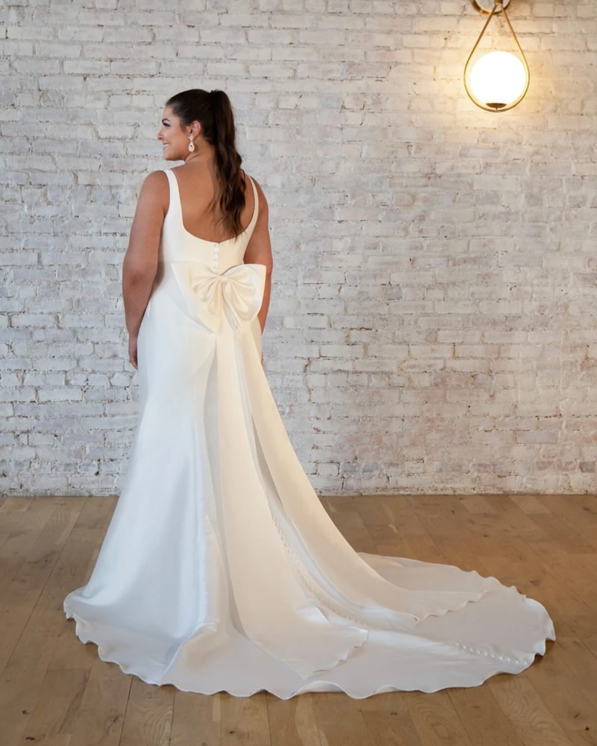 Bride in white gown with bow, turning her head; against a brick wall.