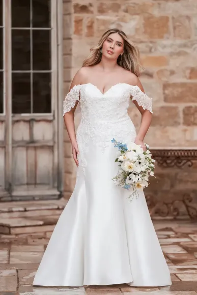 Bride in white off-the-shoulder wedding dress, holding bouquet, standing outside a building.
