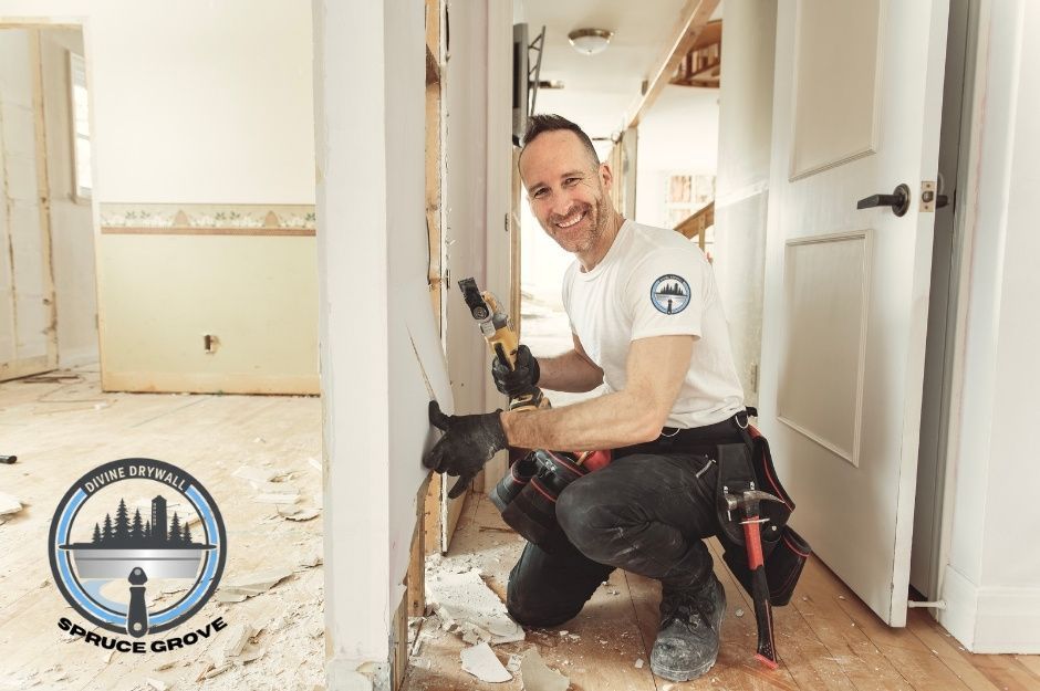 A drywall repair company worker, kneeling on a protective floor covering, holds a power drill while working on baseboards at a commercial construction site. The technician wears a white t-shirt and dark pants in a room with white walls and doorways near 266 St, and 97 Ave, Acheson, AB.