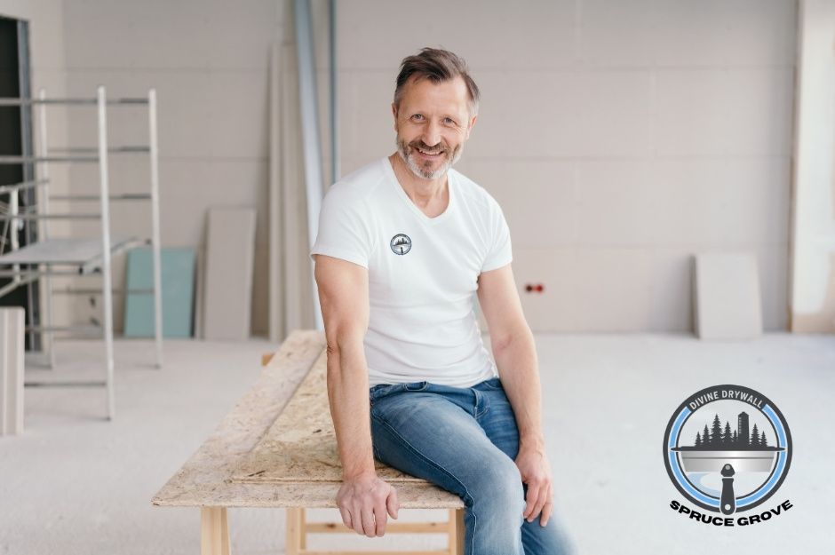 A drywall repair company professional wearing a plain white t-shirt and blue jeans is sitting on a wooden workbench inside a commercial construction site. Behind him are drywall sheets, metal frames, and a scaffold, showing work in progress. Located near 49 St, and 32 Ave, Beaumont, AB.