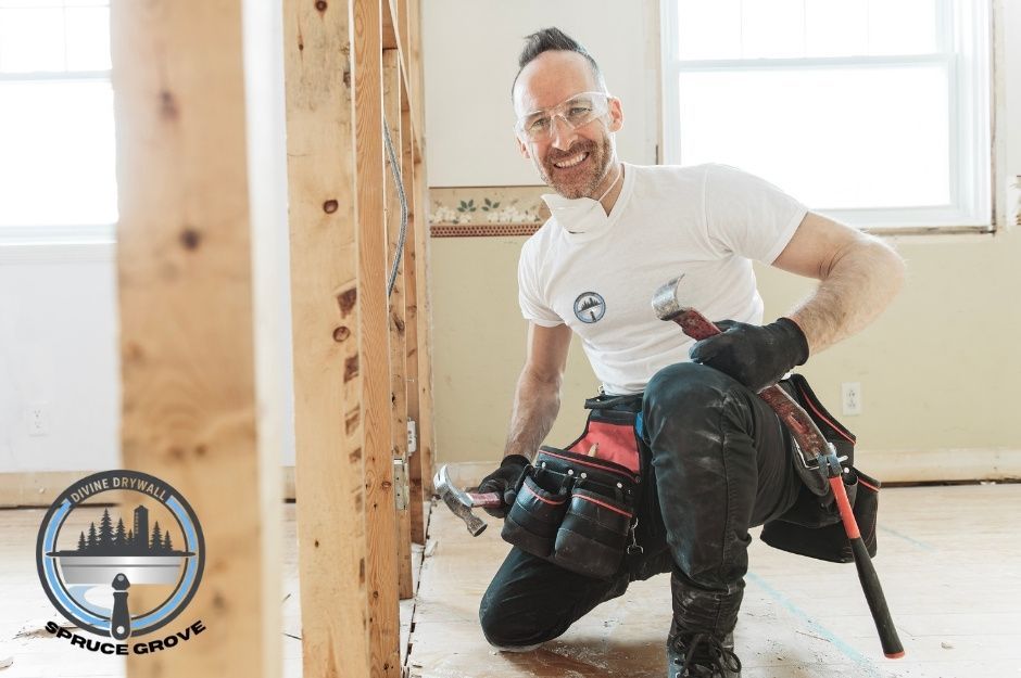 A drywall repair company worker, wearing a white t-shirt and black pants, kneels while holding a hammer in an unfinished commercial area with wooden framing and windows visible. He is working near Oakland Pl, and Highwood Blvd, Devon, AB.