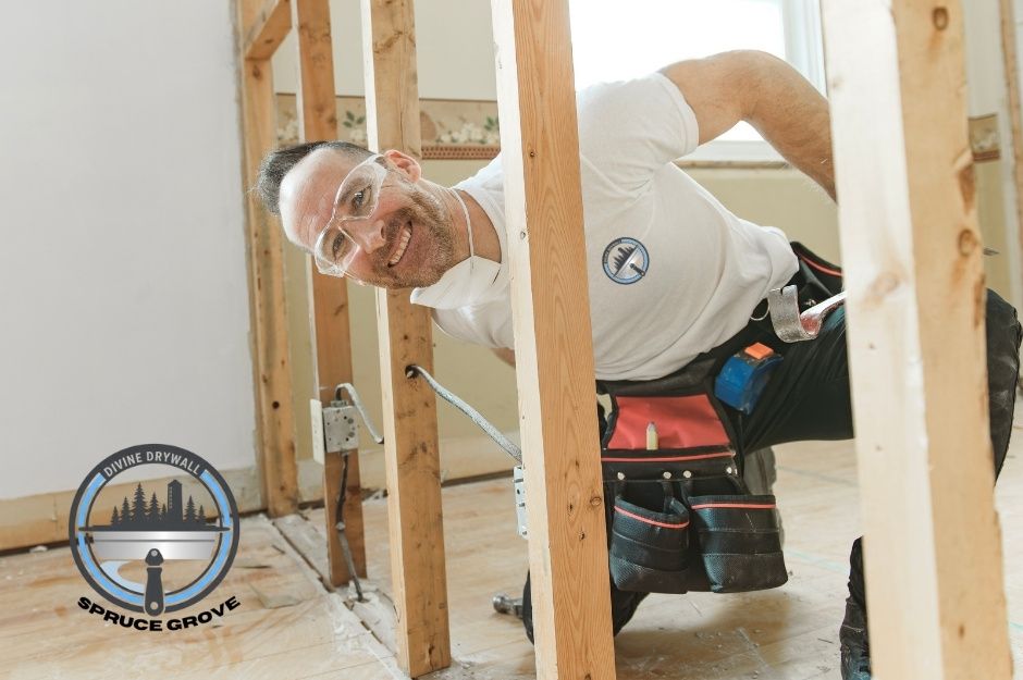 A drywall repair contractor in a white shirt is kneeling behind wooden wall studs, working on a commercial construction project. The contractor performs renovation work near Nootka Rd, and Bella Coola Dr, Leduc, AB.