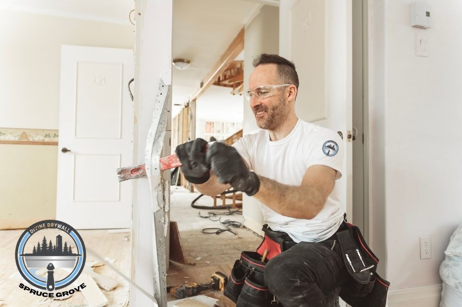 A drywall repair contractor wearing safety glasses, gloves, and a tool belt is using a red pry bar to remove damaged drywall and metal corner bead inside a residential home under renovation. Broken pieces of drywall and tools are scattered on the floor near Briarwood Way, and Graybriar Dr, Stony Plain, AB.