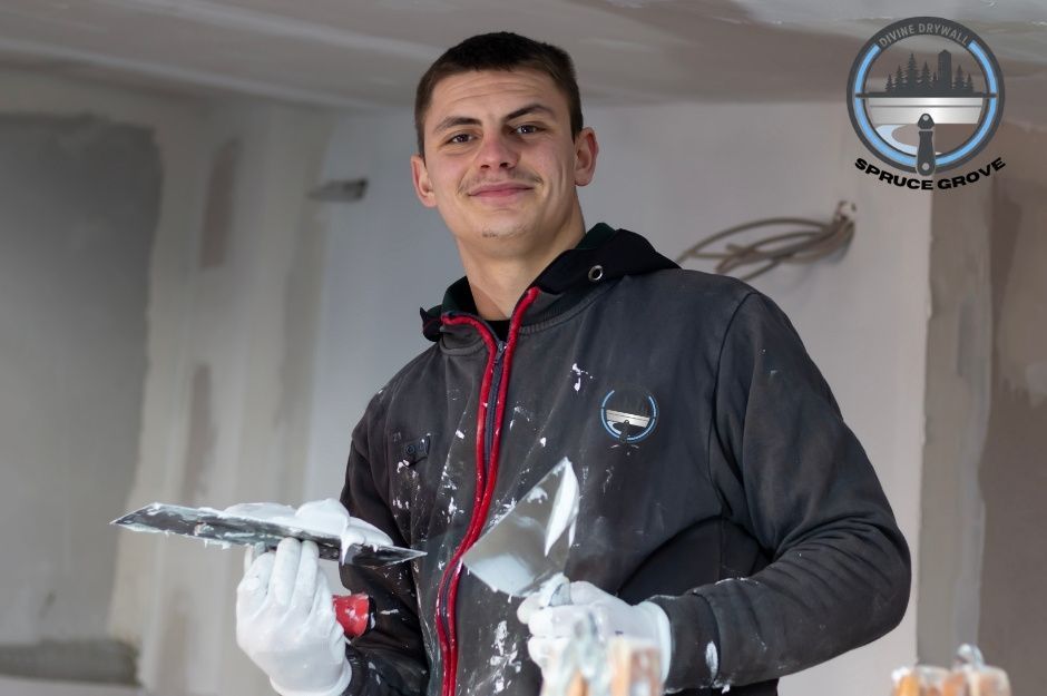 A drywall repair handyman in a black hoodie is smiling while holding a drywall taping knife in one hand and a putty knife in the other, both covered in joint compound. He is working inside a commercial building with unfinished drywall. Located near 53 St, and 38 Ave, Gibbons, AB.