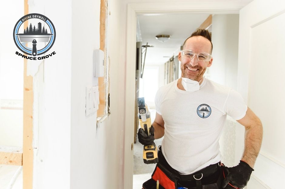 A drywall repair specialist in a white t-shirt smiles while holding a power drill, standing in a commercial hallway with white walls and wooden framing visible near 44 Ave, and 51 St, Calmar, AB.