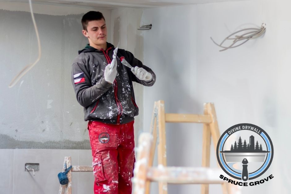 A drywall repair specialist in paint-splattered red pants and a dark hoodie mixes drywall compound using putty knives inside a partially finished white room with exposed wiring and a wooden ladder nearby during a residential project near 99 Ave, and Grandin Dr, Morinville, AB.