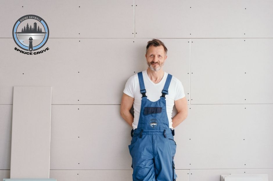 A drywall repair specialist wearing a white t-shirt and blue overalls stands in front of a newly installed drywall surface with visible screw placements. He smiles with his hands behind his back in a commercial project setting. Located near 10 St, and 14 Ave, Nisku, AB.