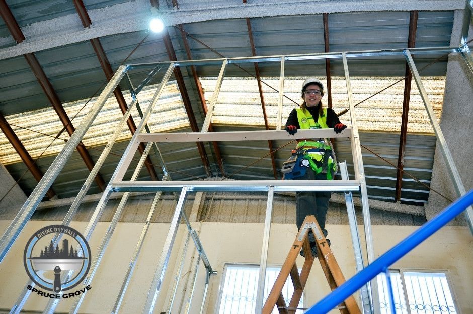 A drywall repair specialist stands on a wooden ladder inside a commercial building, wearing a safety helmet, vest, gloves, and a tool belt, securing a metal frame for drywall installation. Overhead lights and steel beams are visible. Located near Summercourt Terrace, and Summercourt Way, Sherwood Park, AB.