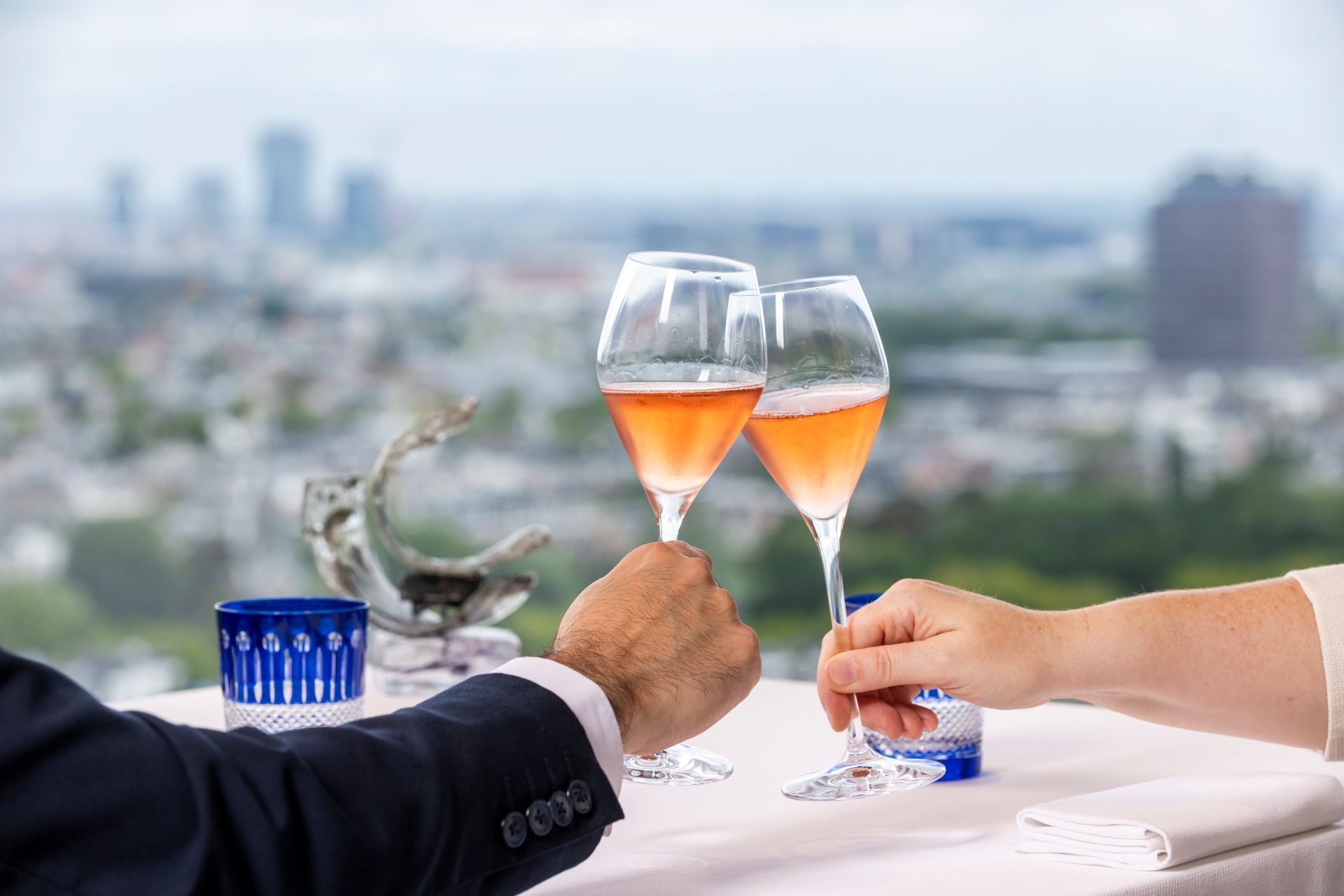 Two people toast with rosé at a rooftop table overlooking a blurred city skyline.