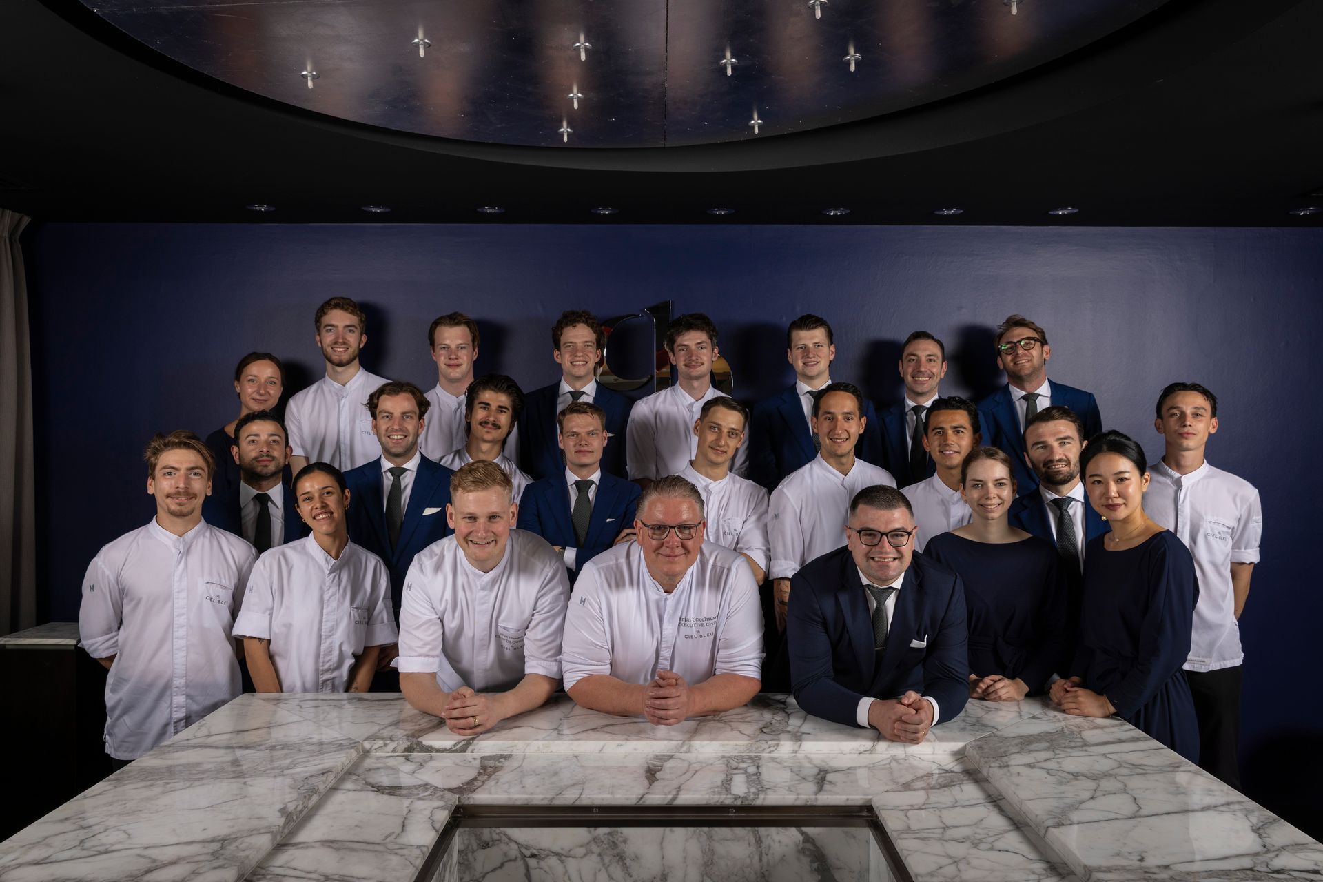Large group of professionals posing in a conference room behind a marble table, with a dark ceiling and wall backdrop