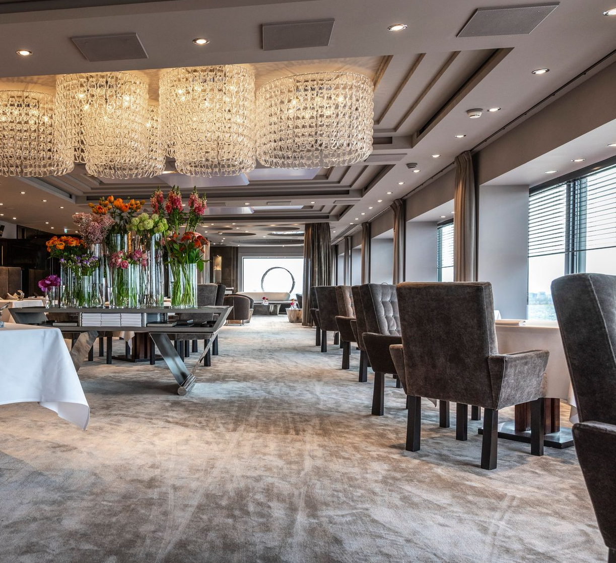Elegant banquet hall with chandeliers, long tables, floral centerpiece, and upholstered chairs by large windows