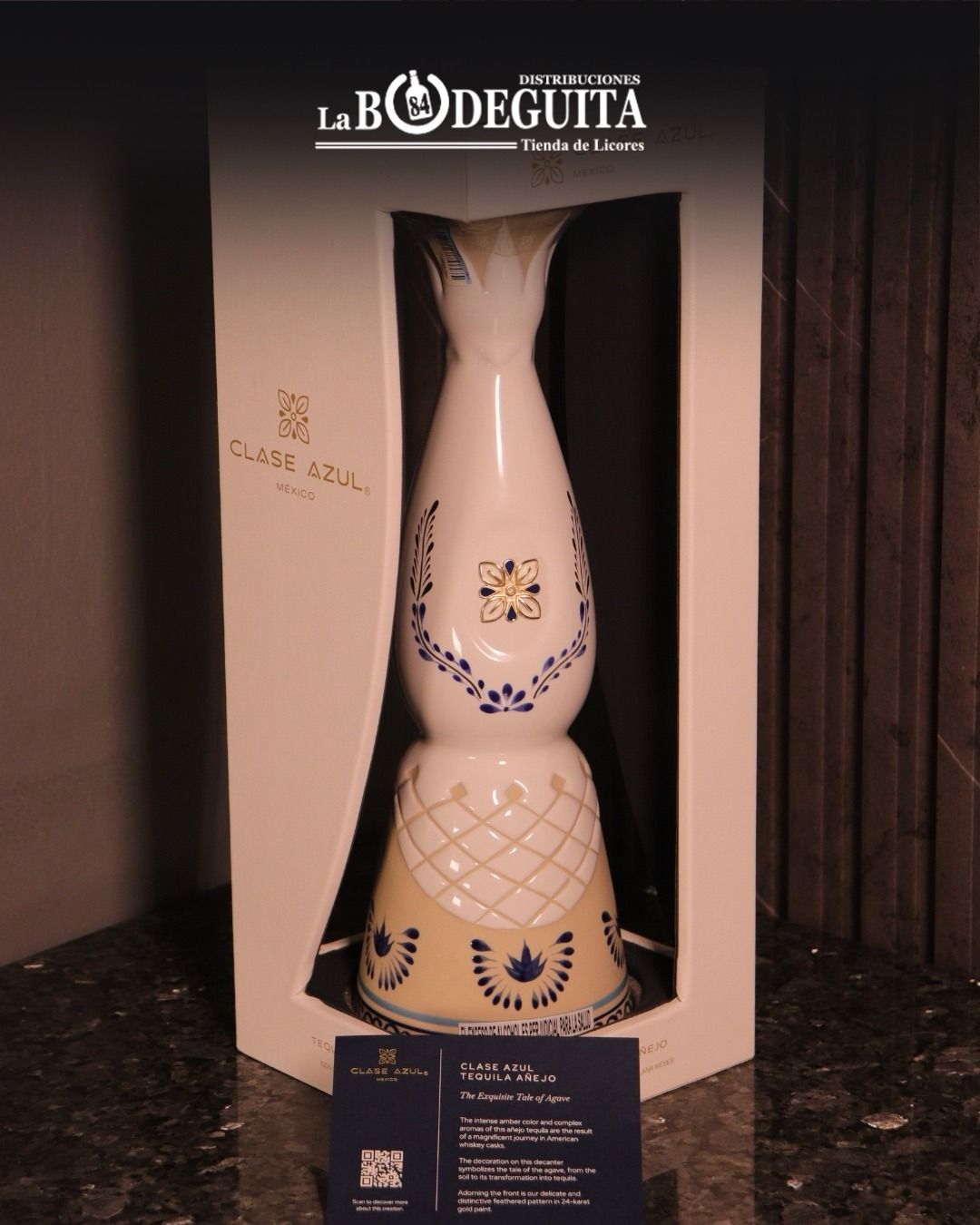Botella de tequila con un diseño decorativo en blanco y azul, dentro de una caja blanca, sobre una superficie oscura.