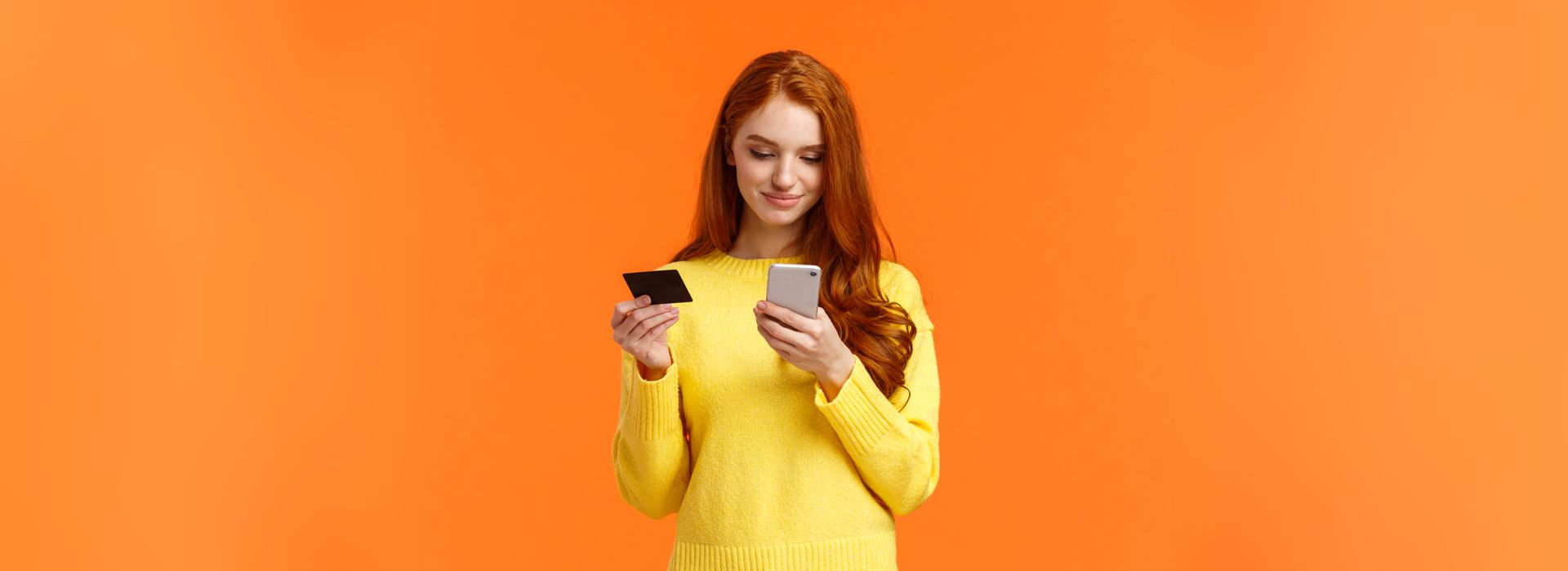 Une femme portant un pull jaune tient une carte de crédit et un téléphone portable.