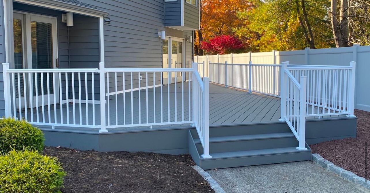 Gray deck with white railing and stairs leading to the house. Autumn trees in the background.