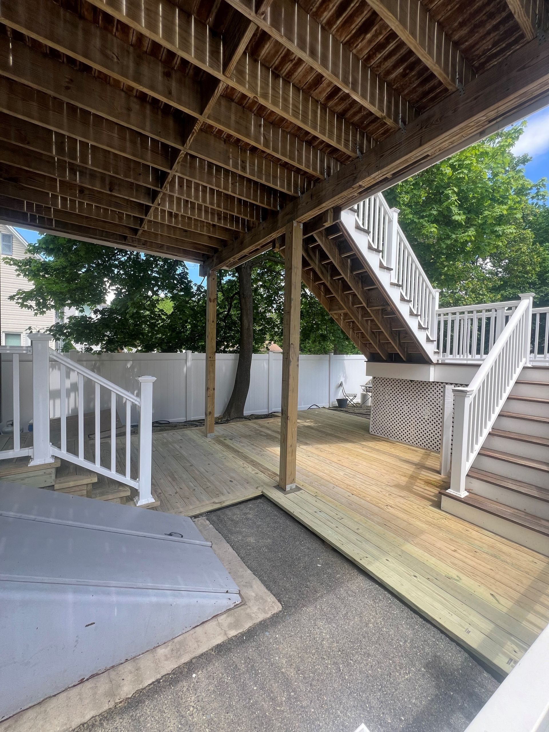 Wooden deck with stairs, railings, and supporting beams. White fence and trees in the background.