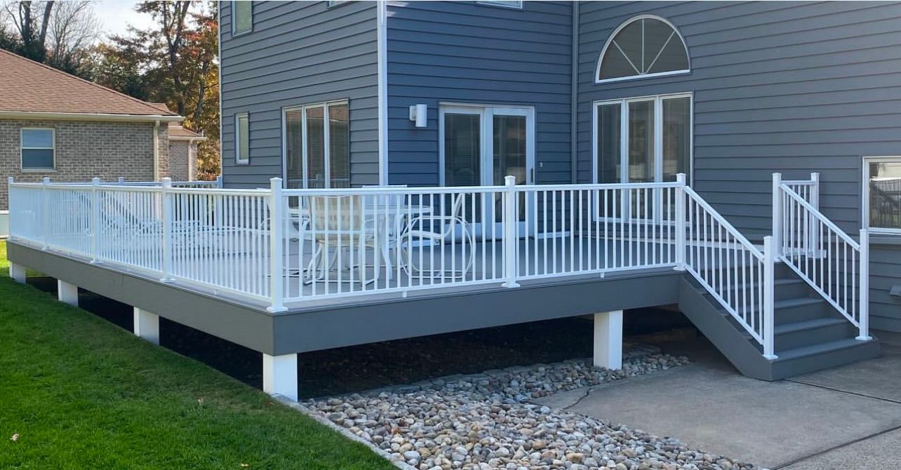 A gray deck with white railings and stairs extending from a gray house with a lawn and stone path.