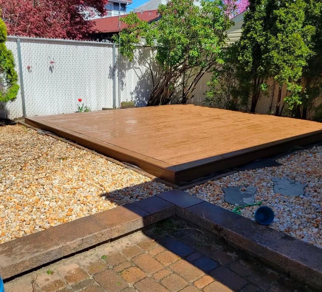 Raised wooden platform filled with tan sand in a backyard setting, surrounded by gravel and landscaping.