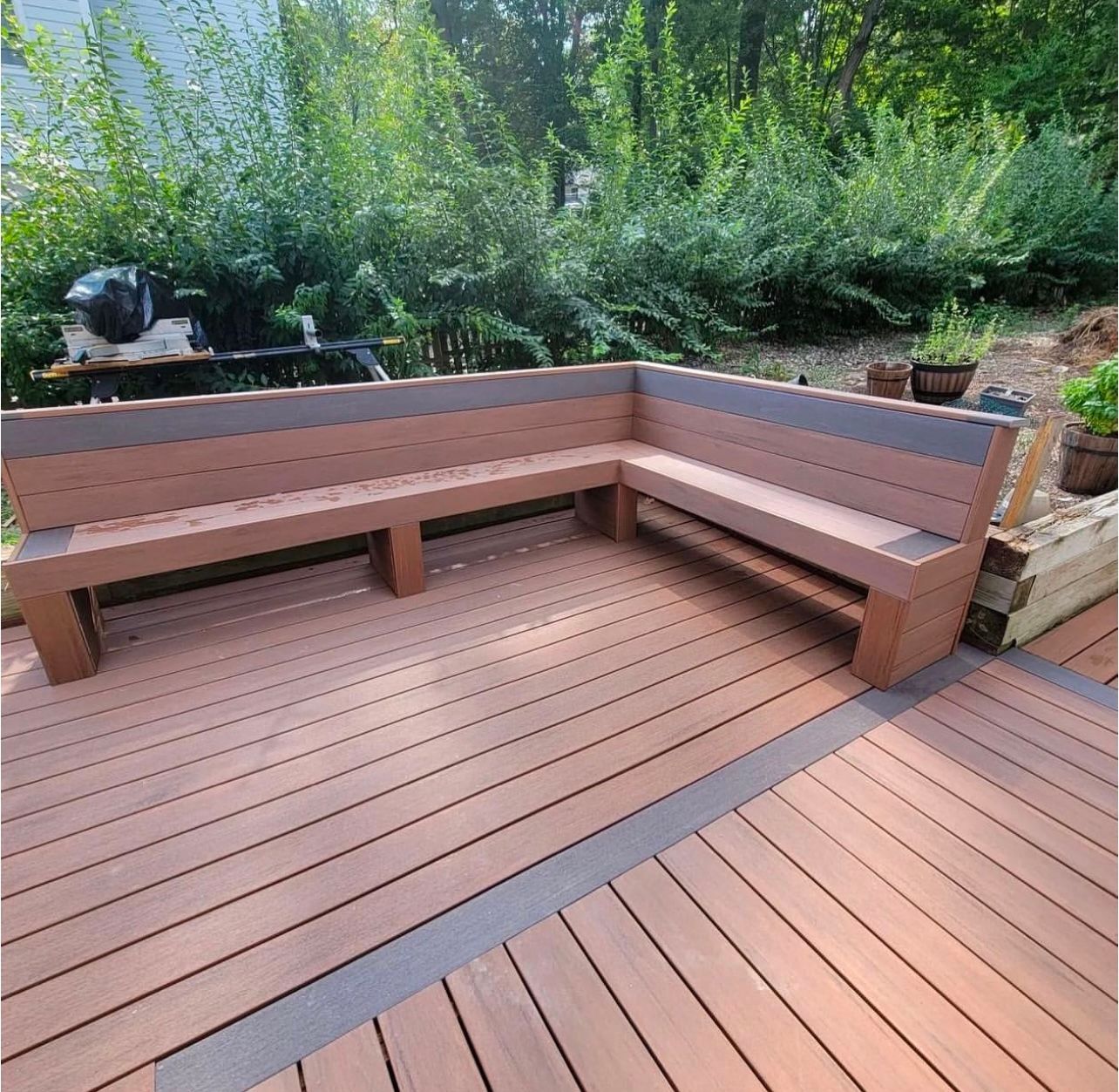 L-shaped brown and gray bench on a wooden deck, surrounded by greenery.
