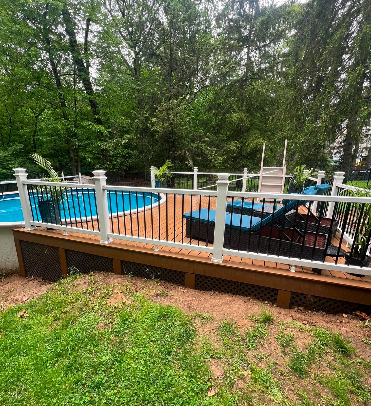 Above-ground pool with a wooden deck and white railings, surrounded by trees and grass.