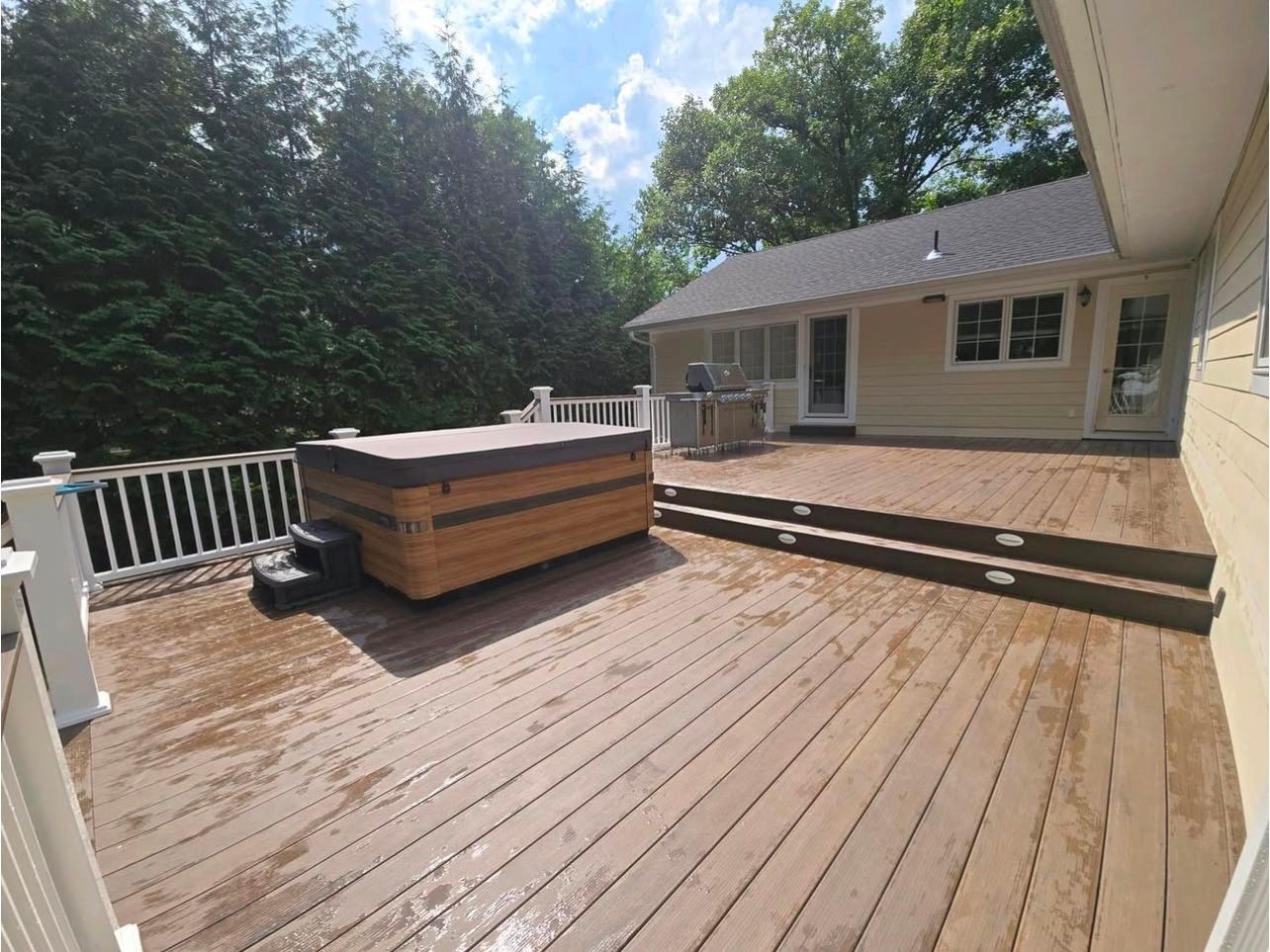 A wooden deck with a hot tub, leading to a light yellow house with a green tree background on a sunny day.