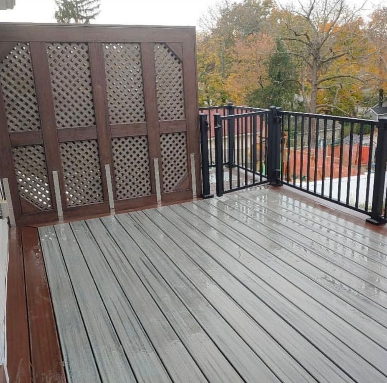 Wet outdoor deck with brown latticed privacy screen and black railing; fall foliage in background.