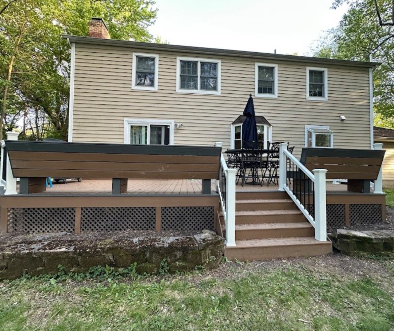 Back of a two-story house with a deck. Brown deck with benches, stairs, and outdoor furniture. Green lawn surrounds.